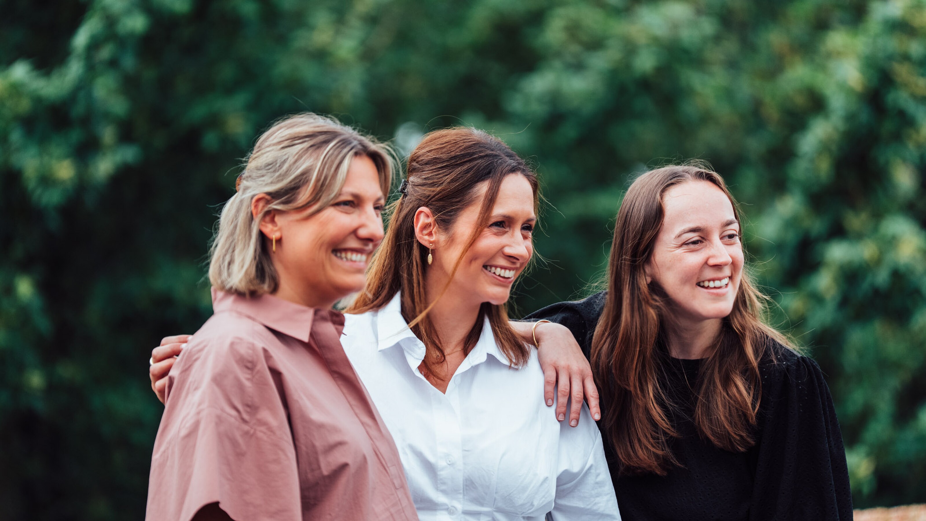Drie vrouwen glimlachen samen buiten, met groene bomen op de achtergrond.