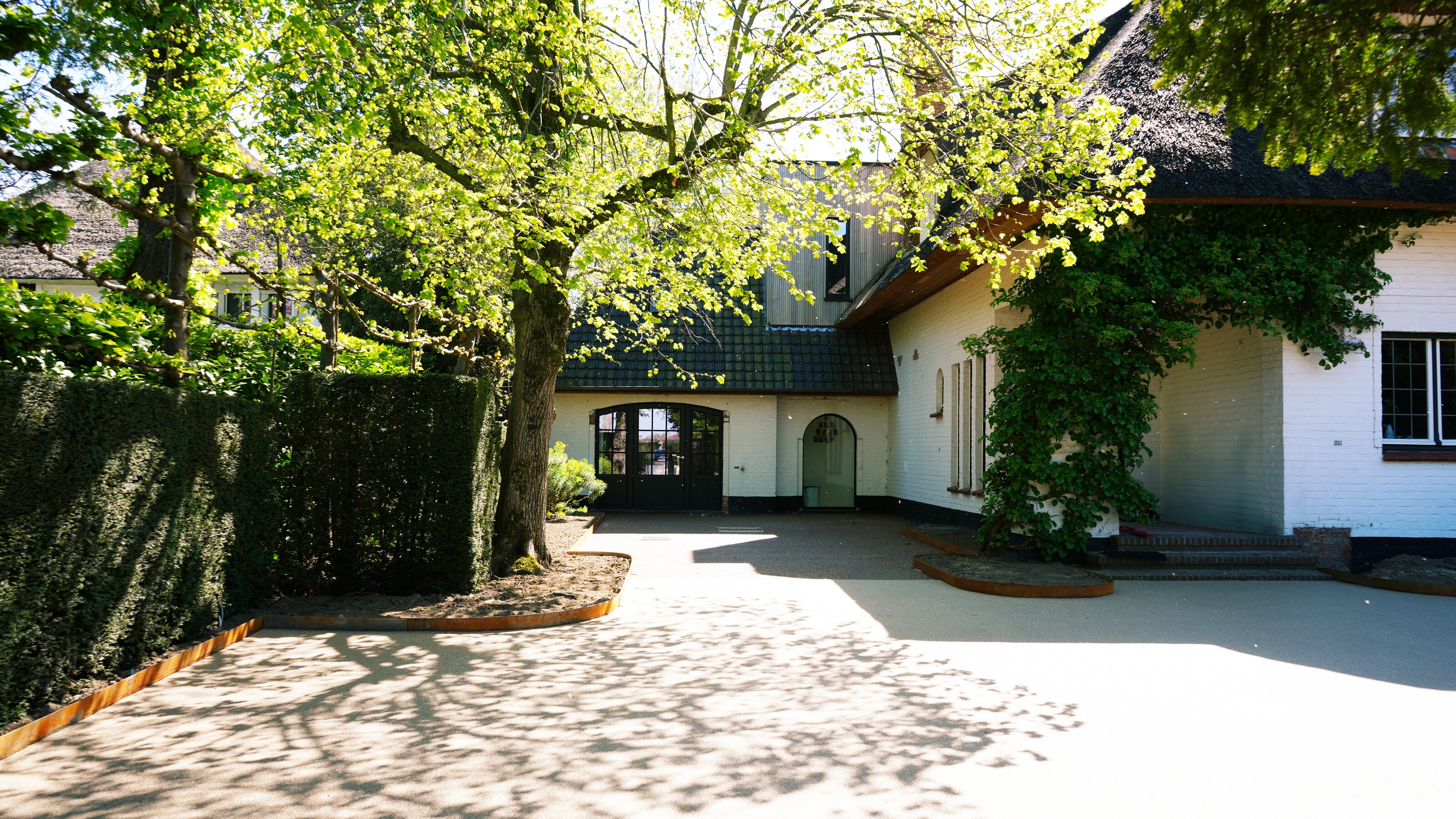 Zonnige oprit met kasseien en schaduw van boom bij wit huis met garage