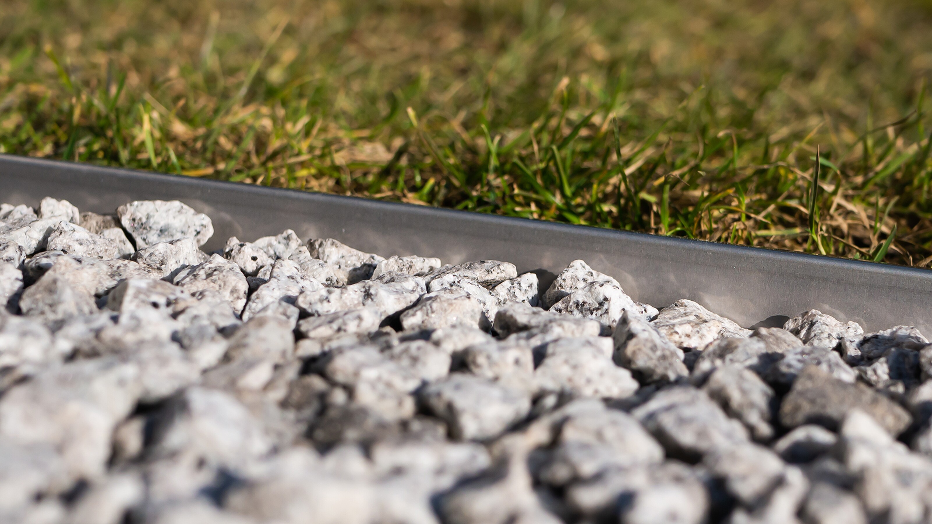 Close-up van grijze grindstenen langs een metalen rand, met gras op de achtergrond.