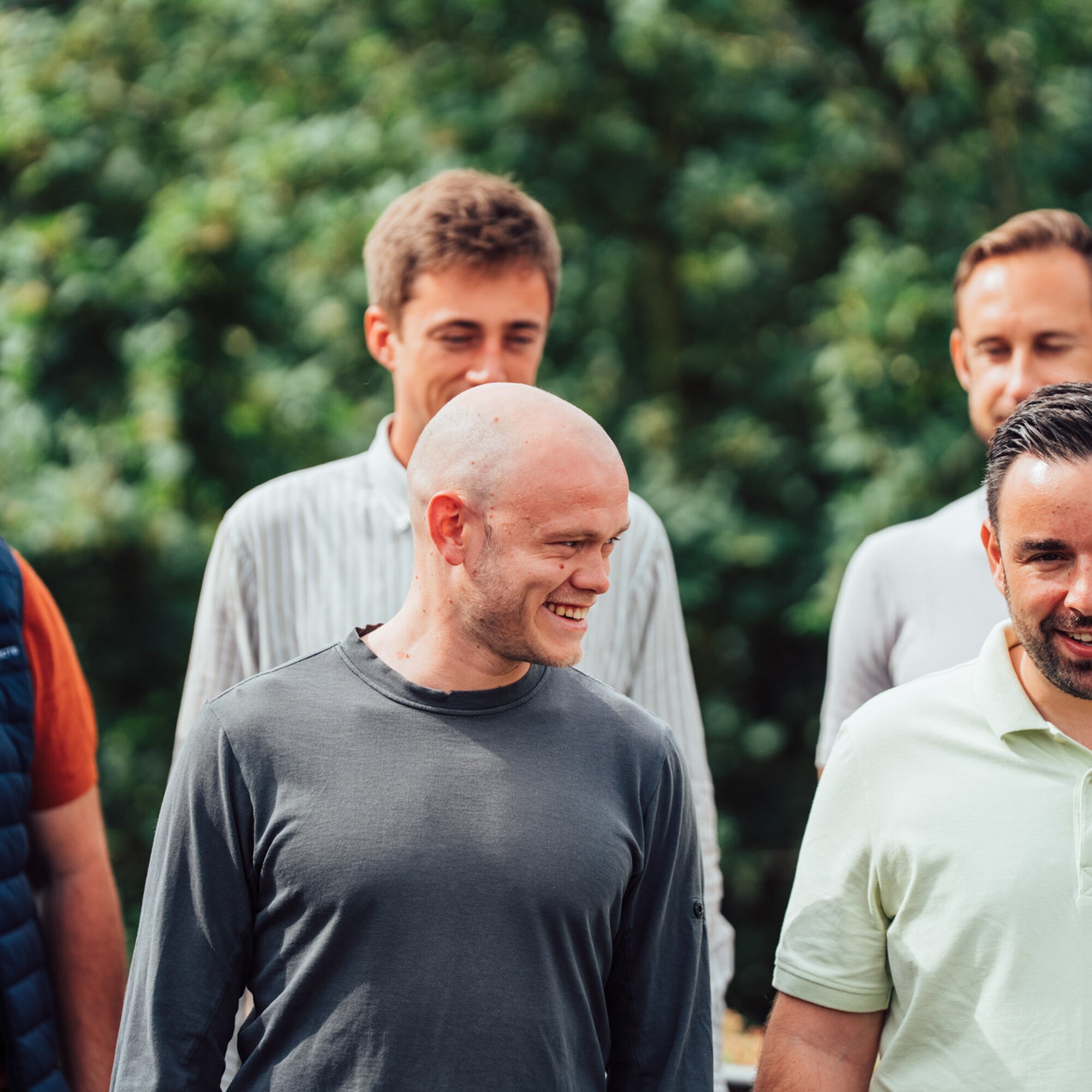 Vijf mannen wandelen buiten; twee lachen, groen gebladerte op de achtergrond.