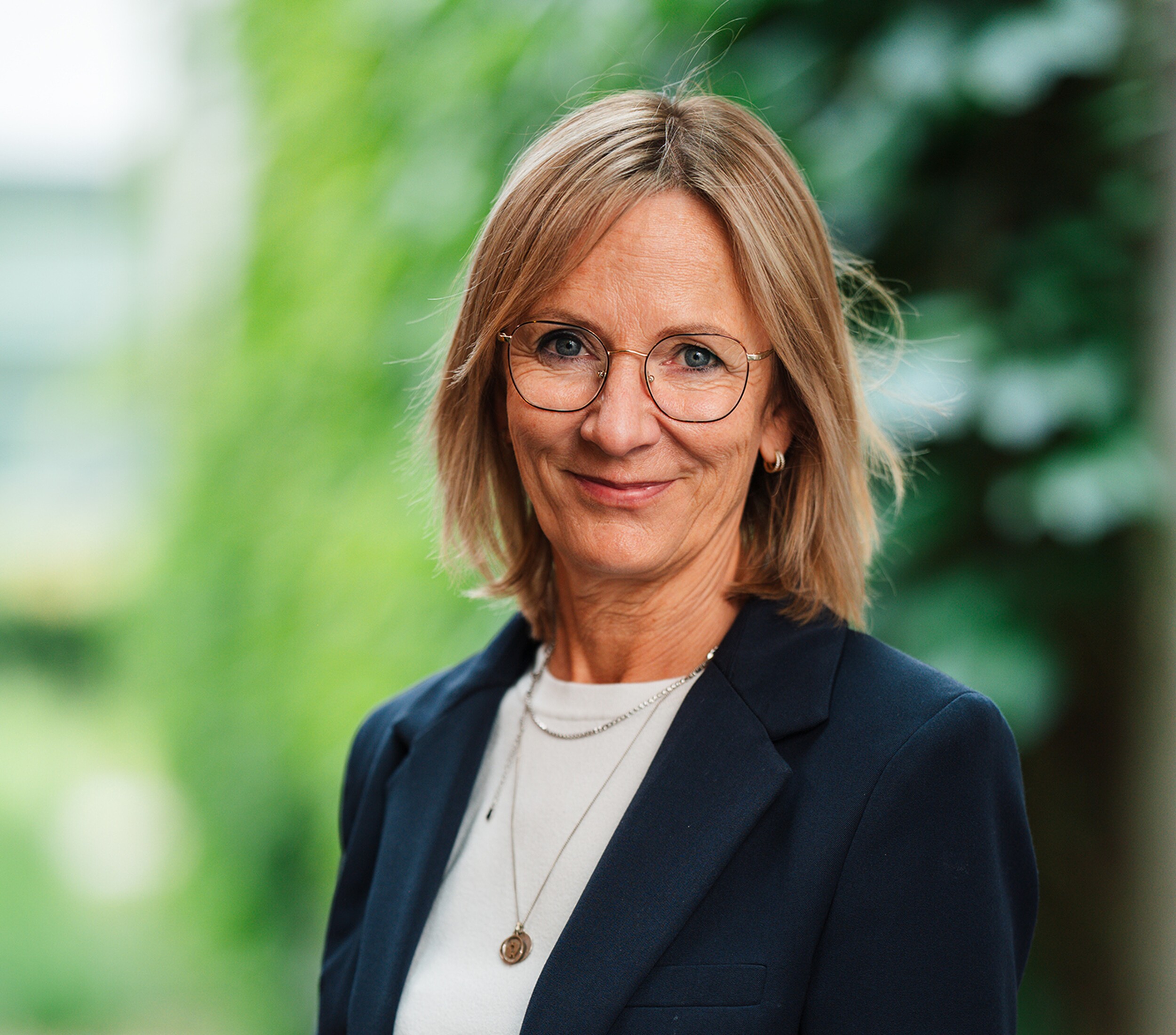 Portretfoto van vrouw met bril en blazer, glimlachend tegen groene achtergrond