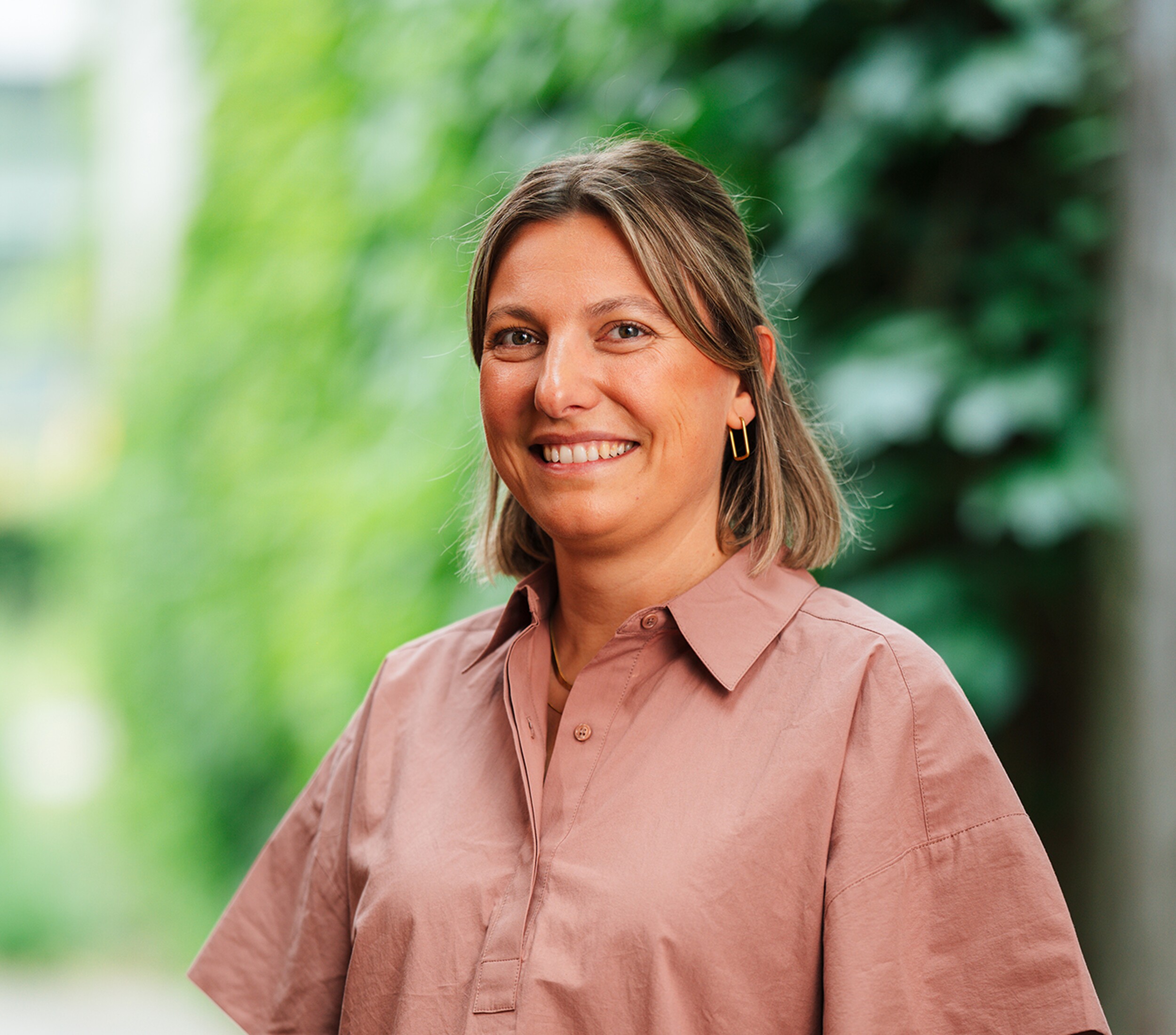 Lachende vrouw met bruin haar in roze blouse, buiten in het groen.