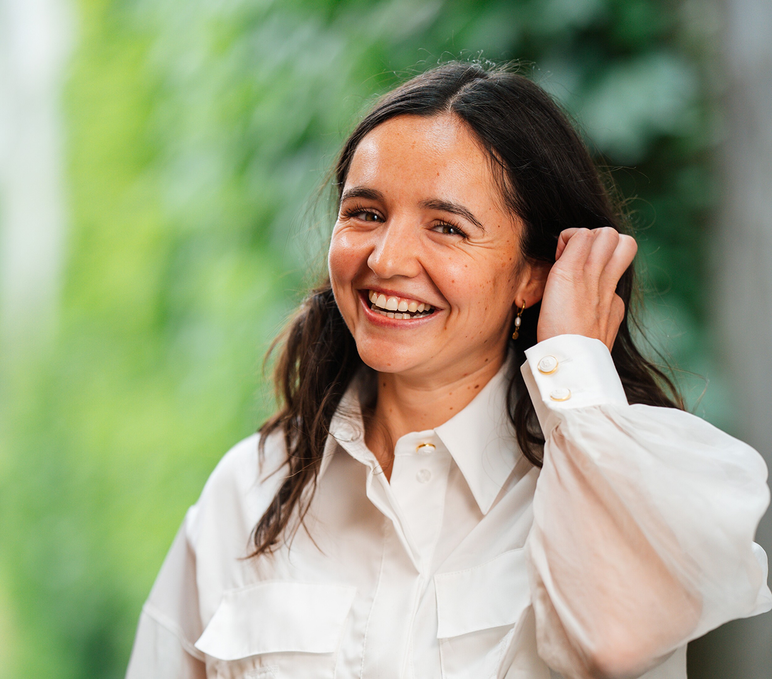 Lachende vrouw in witte blouse, hand in haar haar, groene achtergrond.