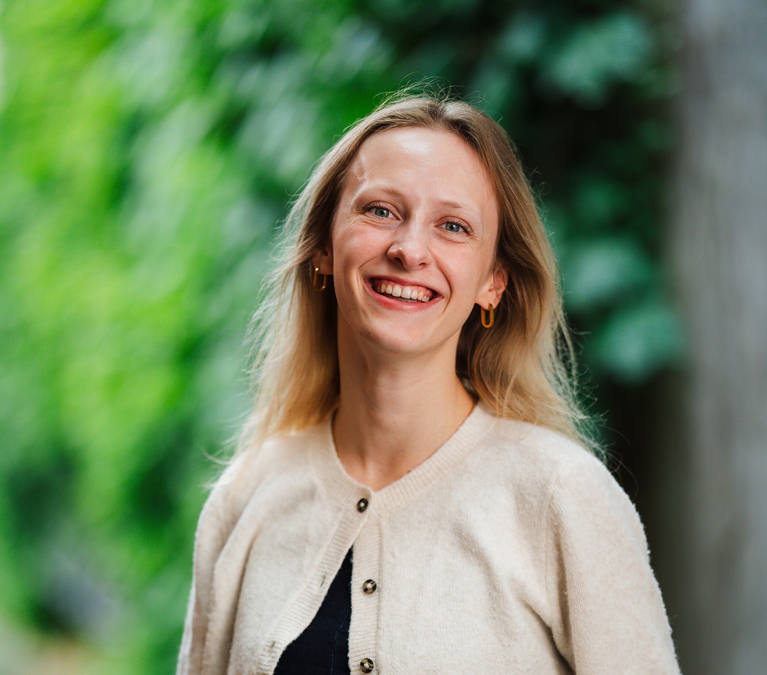 Lachende vrouw met blond haar in beige vest, buiten met groene achtergrond.