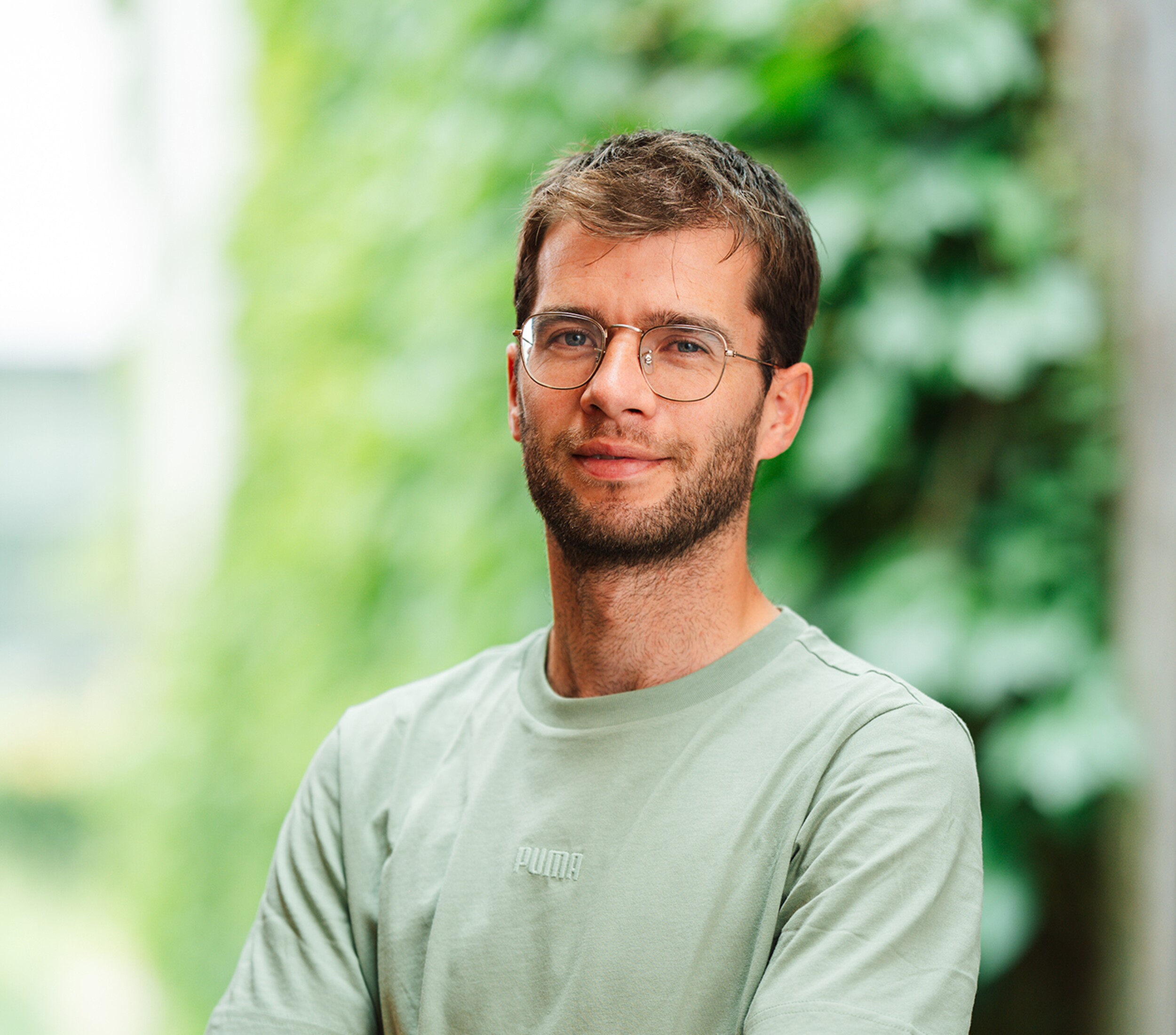 Portretfoto van een man met bril en groene trui, zachte achtergrond van groen blad.