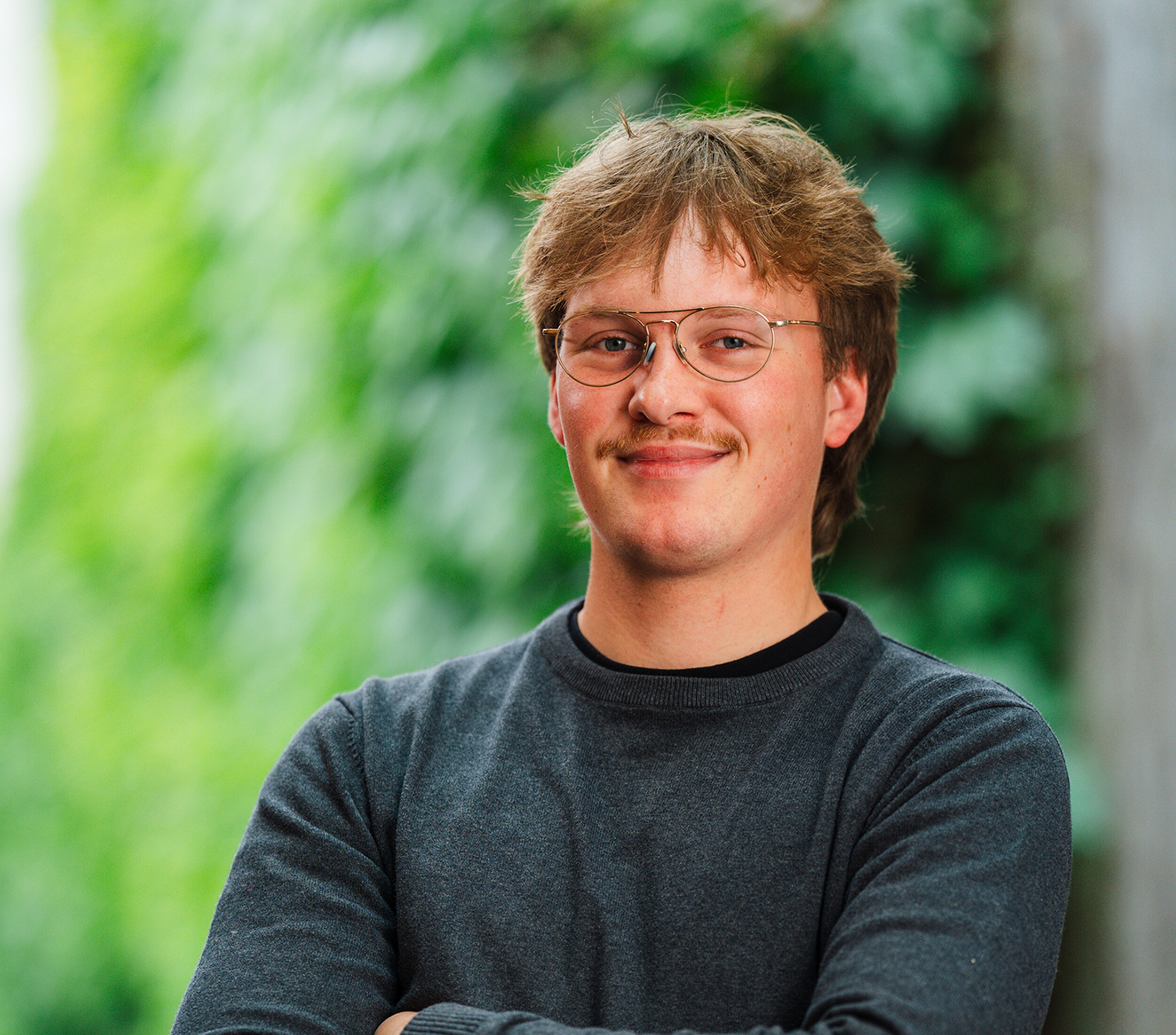 Glimlachende man met bril en gekruiste armen, tegen groene wazige achtergrond.