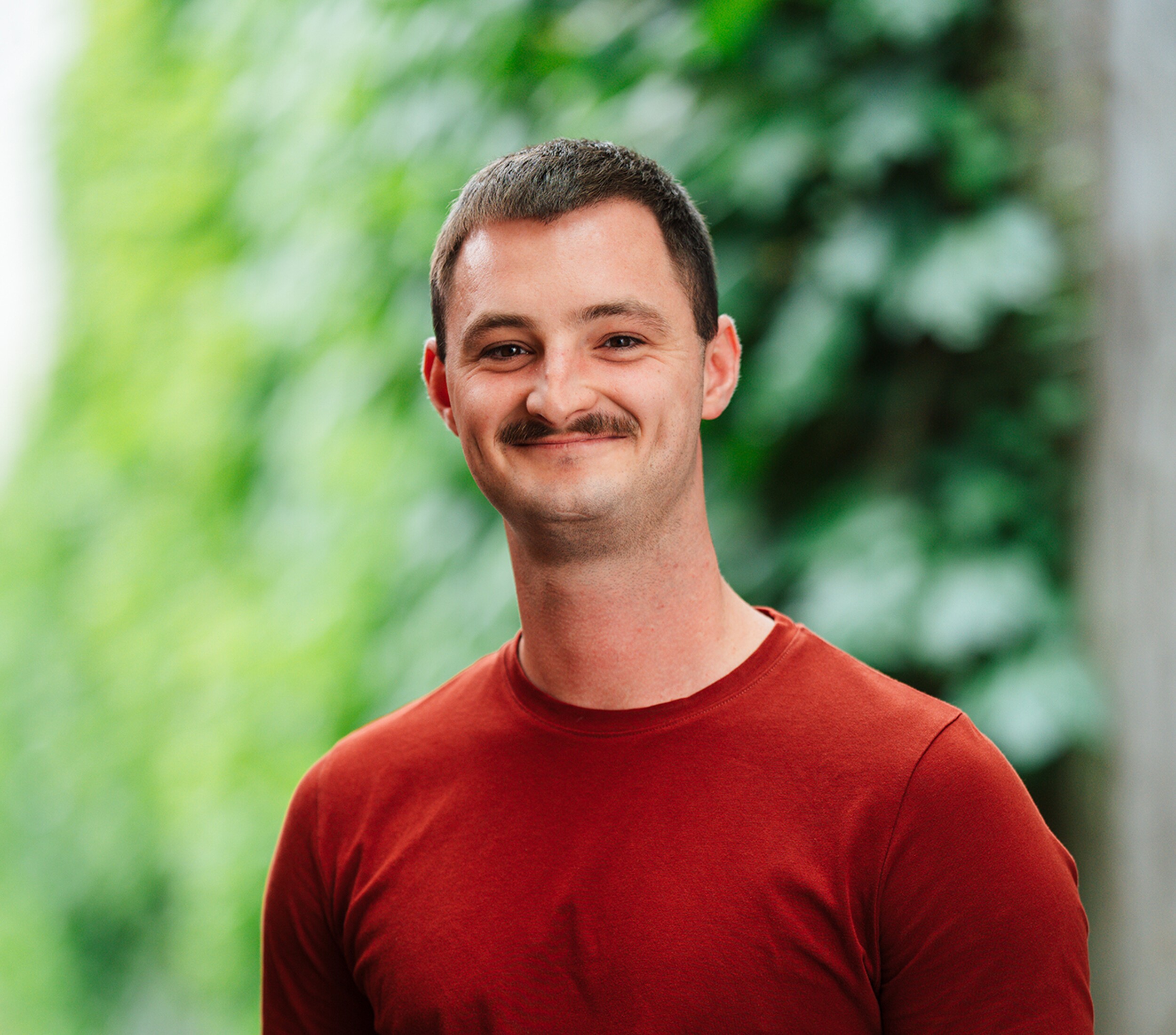 Glimlachende man in rood T-shirt, buiten met groene achtergrond.