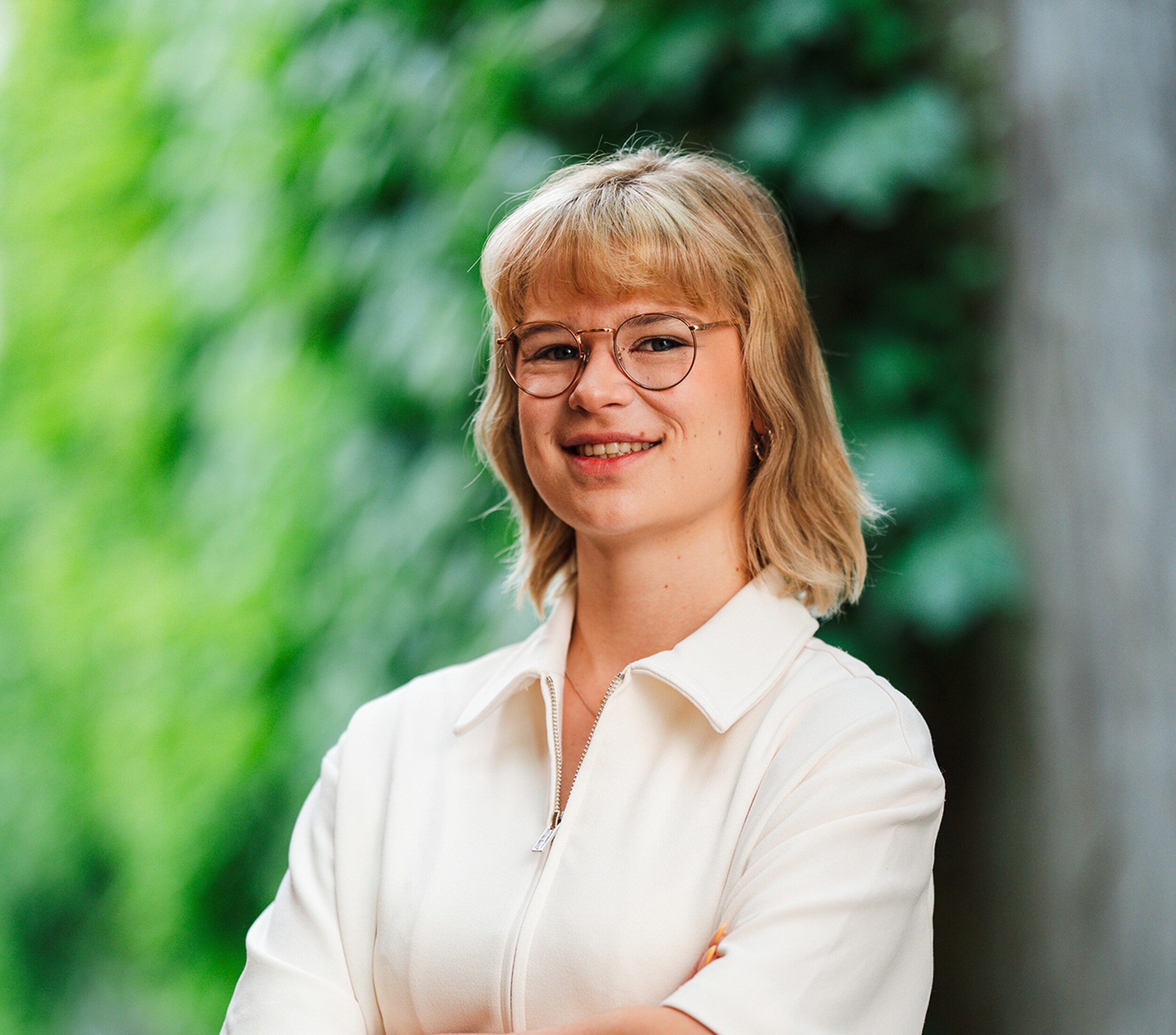 Lachende vrouw met bril en blond haar in witte blouse, groene achtergrond.