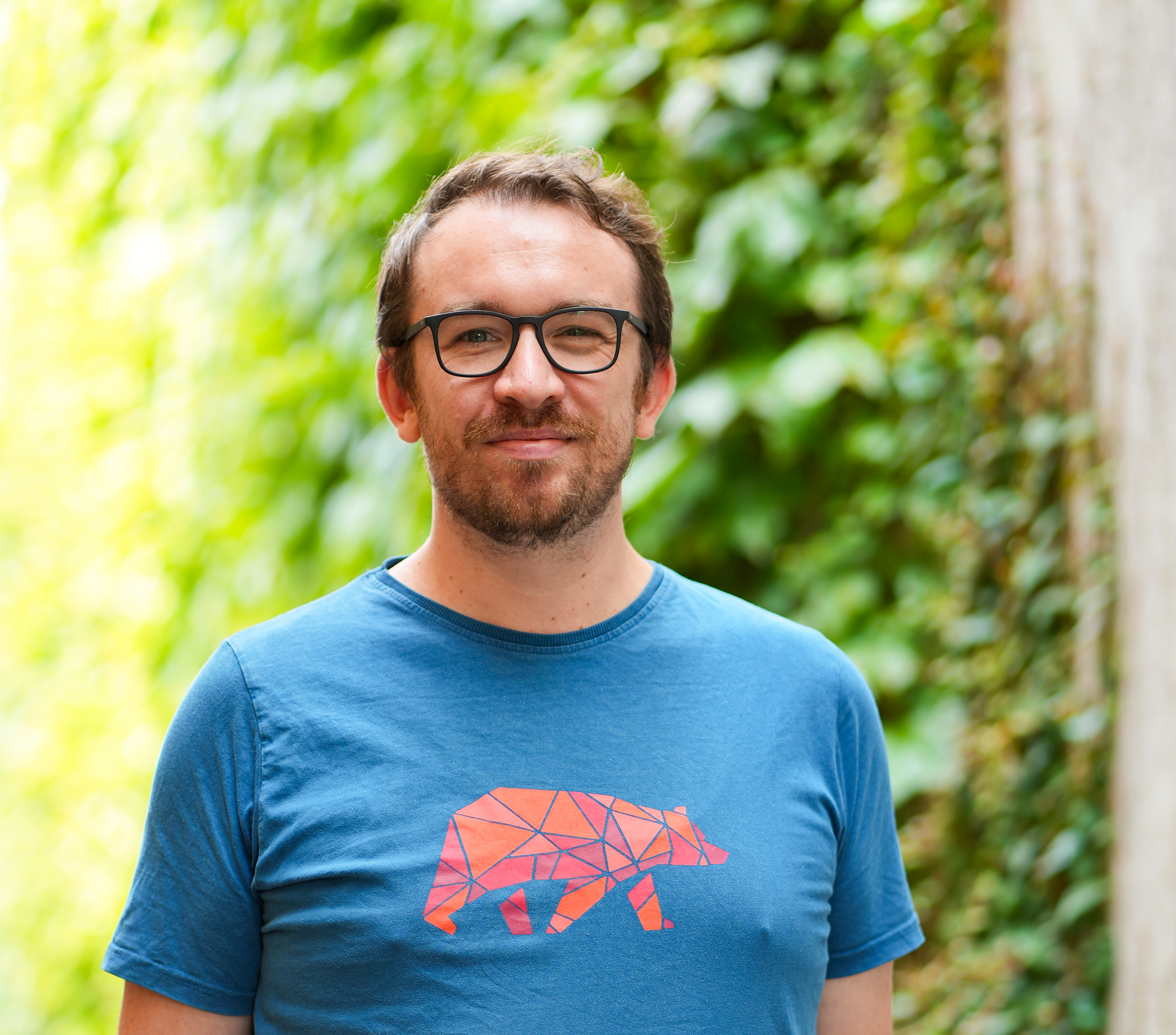Man with glasses in blue T-shirt standing outdoors by a tree and greenery