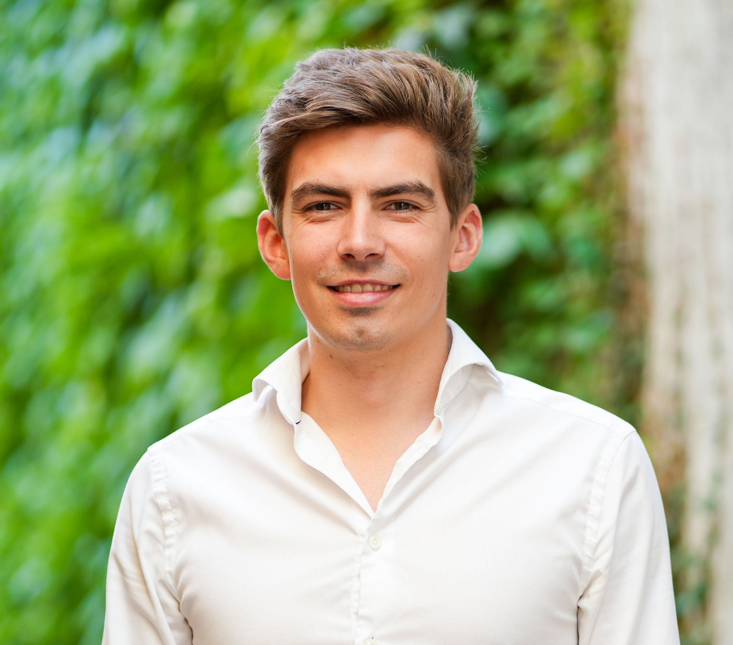 Man in a white shirt smiling outdoors beside a stone wall and greenery