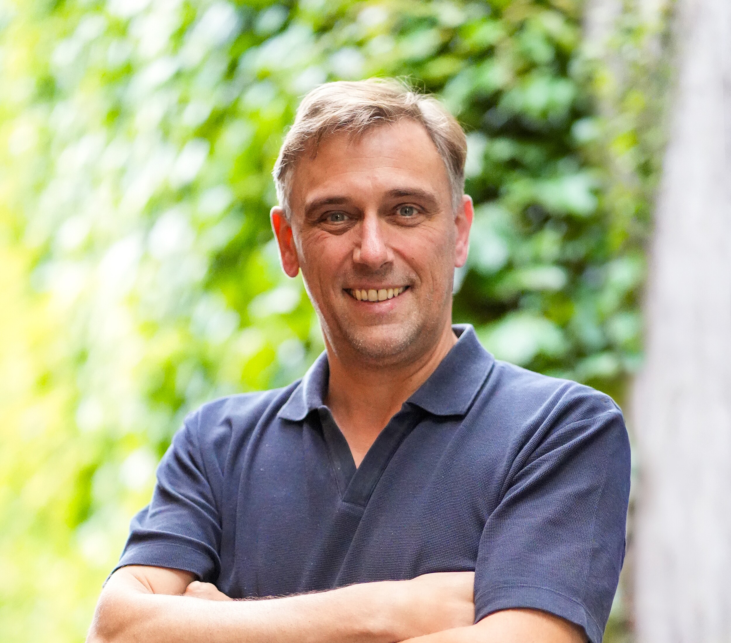 Smiling man with arms crossed, wearing a navy polo in front of green foliage