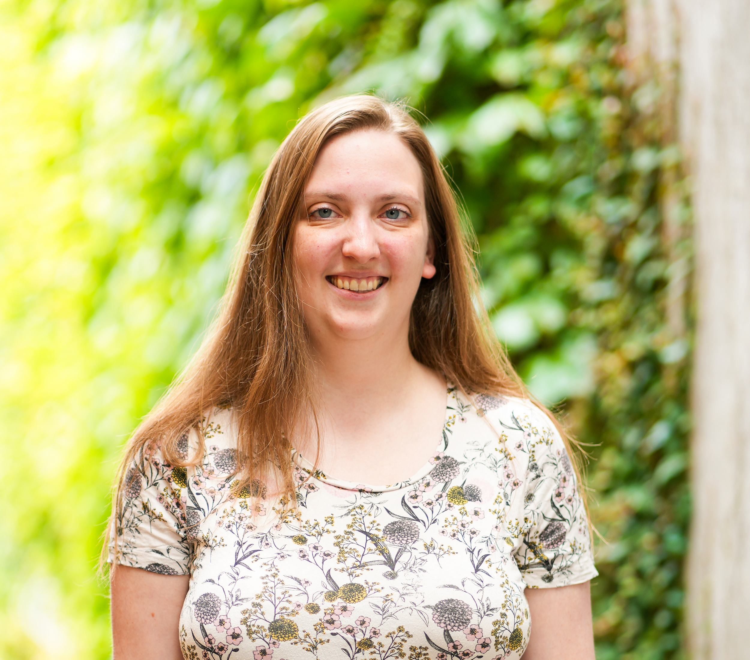 Smiling woman in floral top standing outdoors by a tree with greenery behind