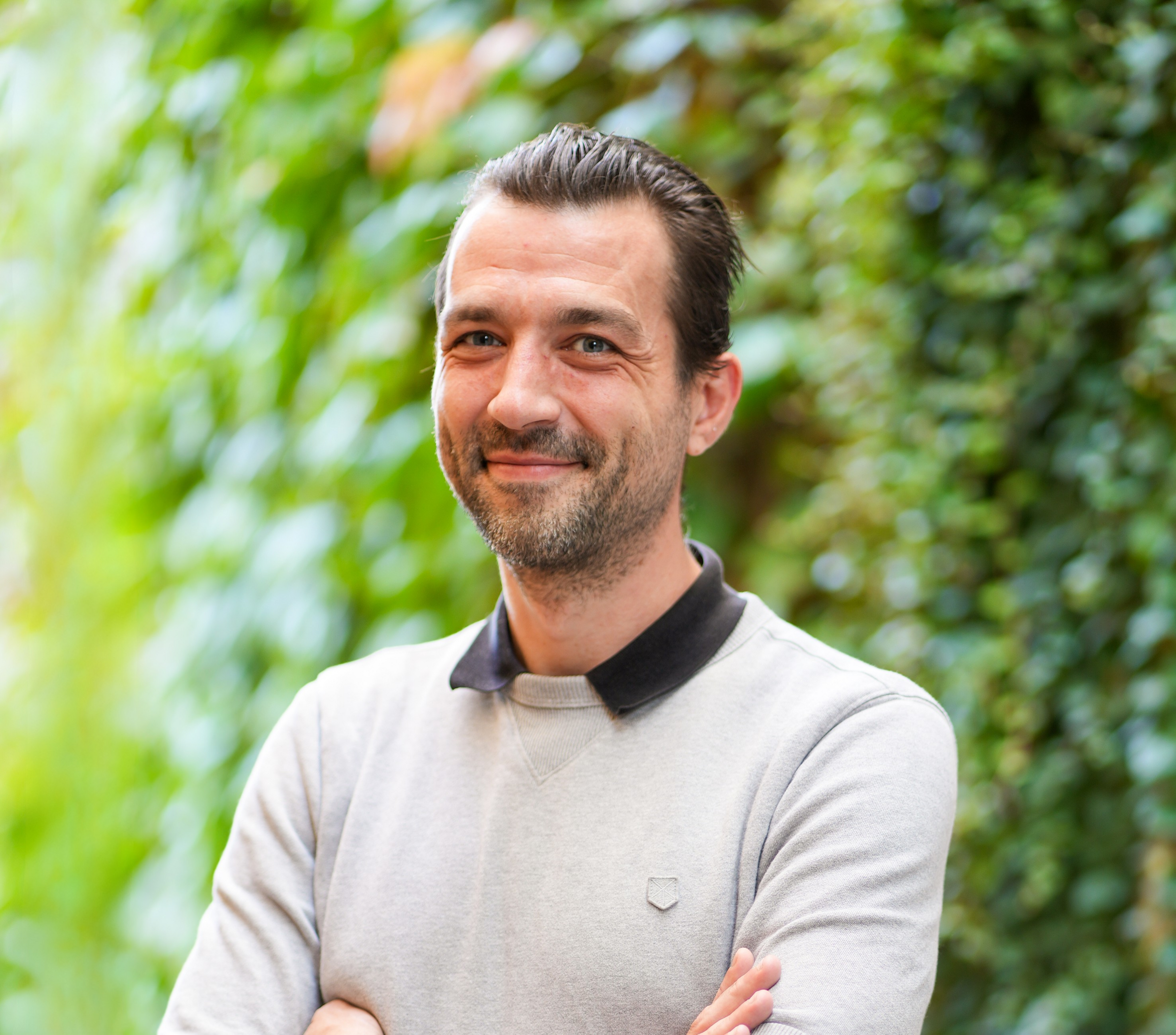 Smiling man with arms crossed, standing against a leafy green wall