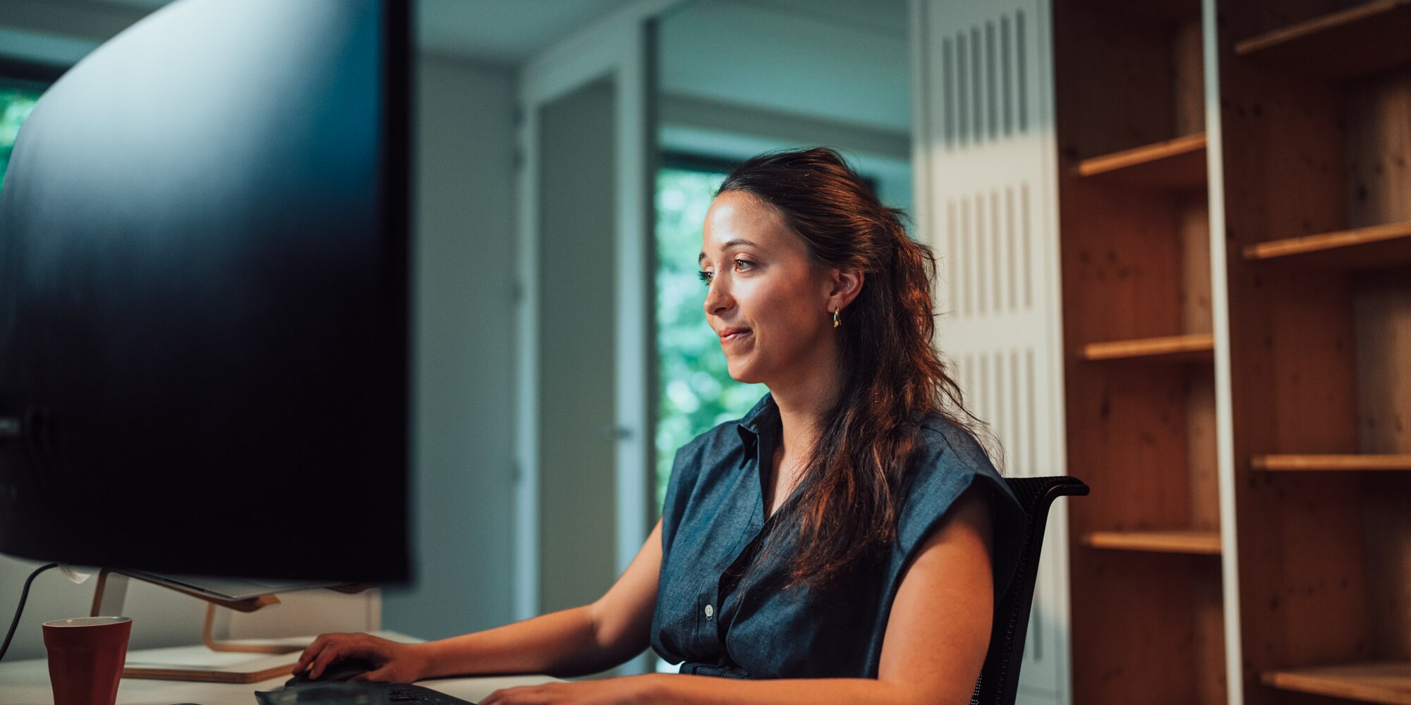 Vrouw werkt aan computer in kantoor, met grote monitor en houten rekken op de achtergrond.