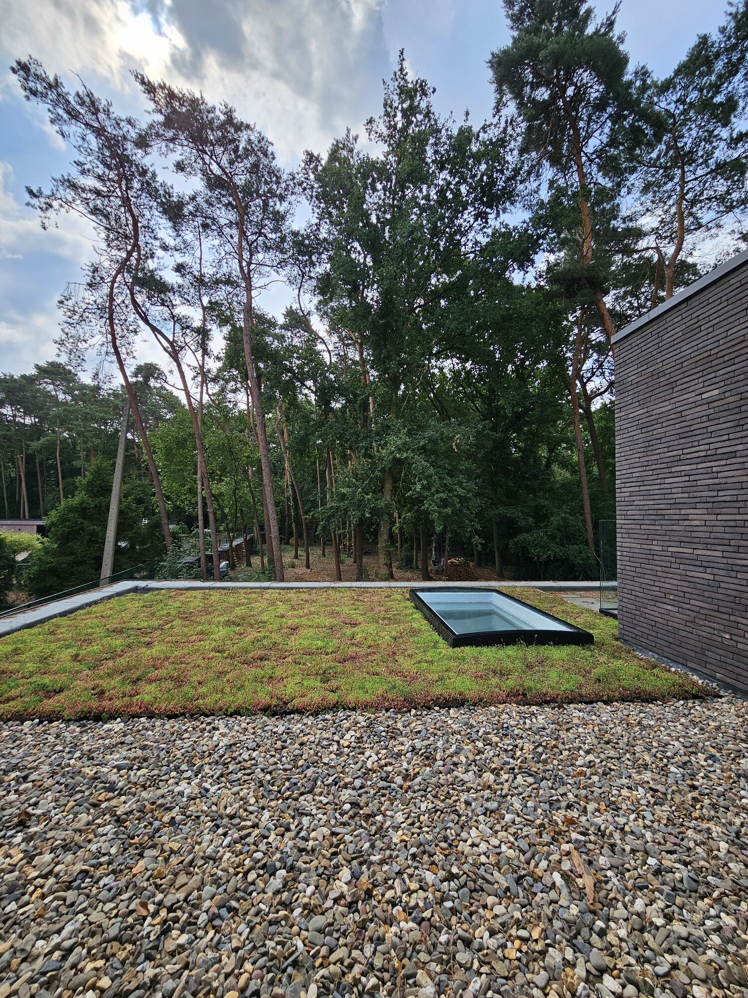 Gravel terrace next to a brick wall, overlooking a lawn with a flat roof skylight and trees.
