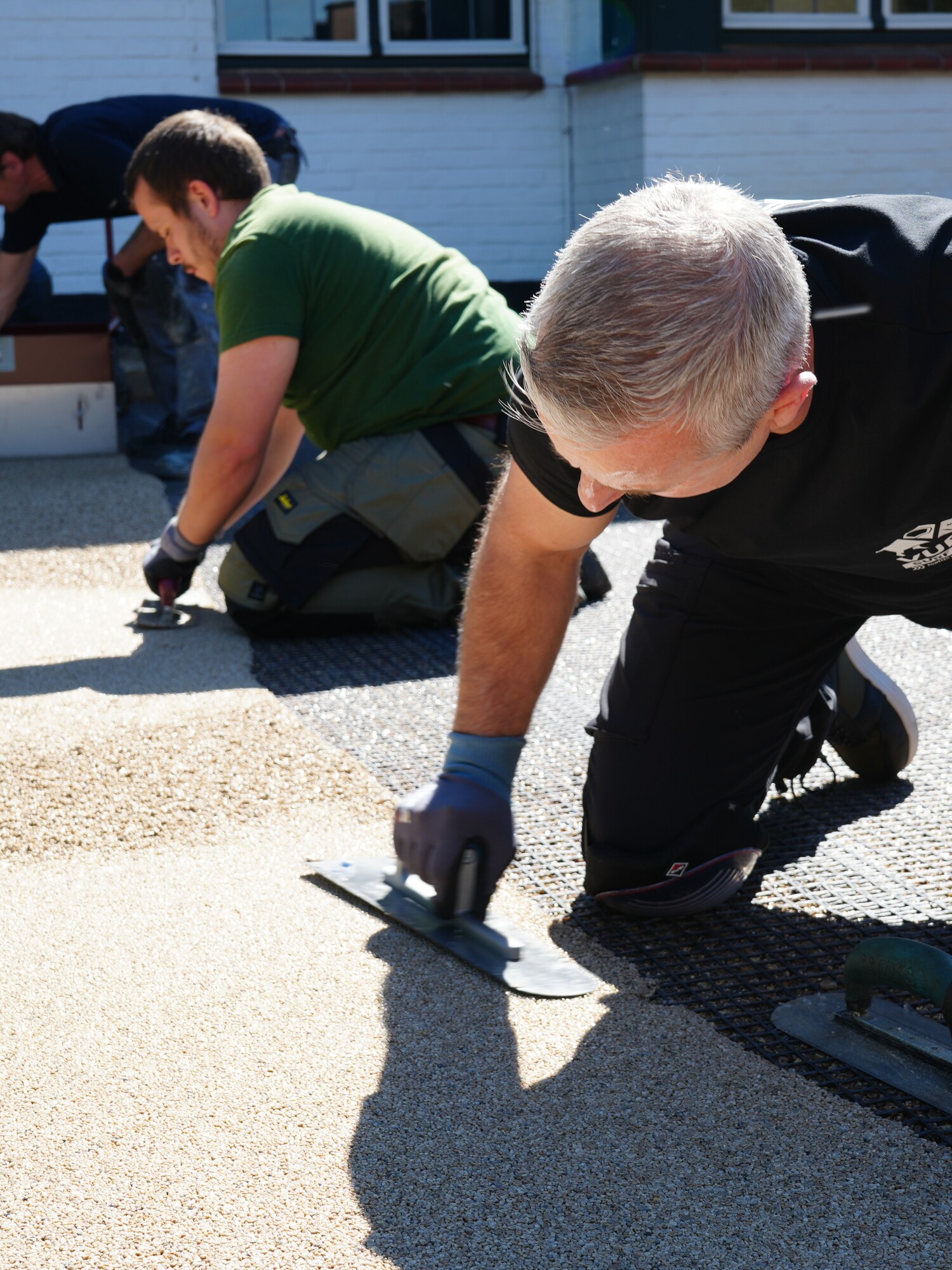 Two people kneeling on pavement, smoothing an asphalt patch outside a building