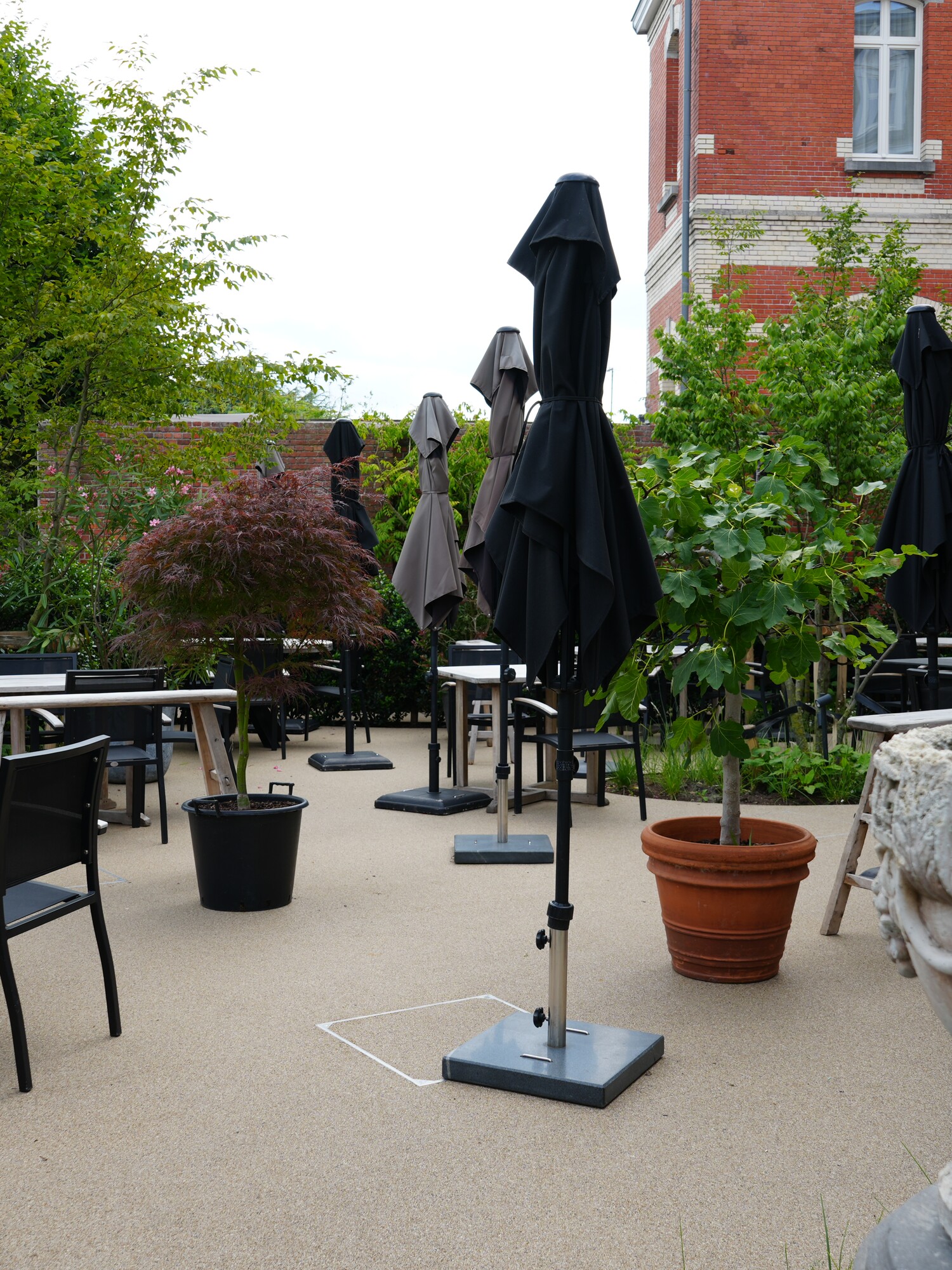 Outdoor terrace with closed umbrellas, empty tables and chairs, plants and a stone vase.