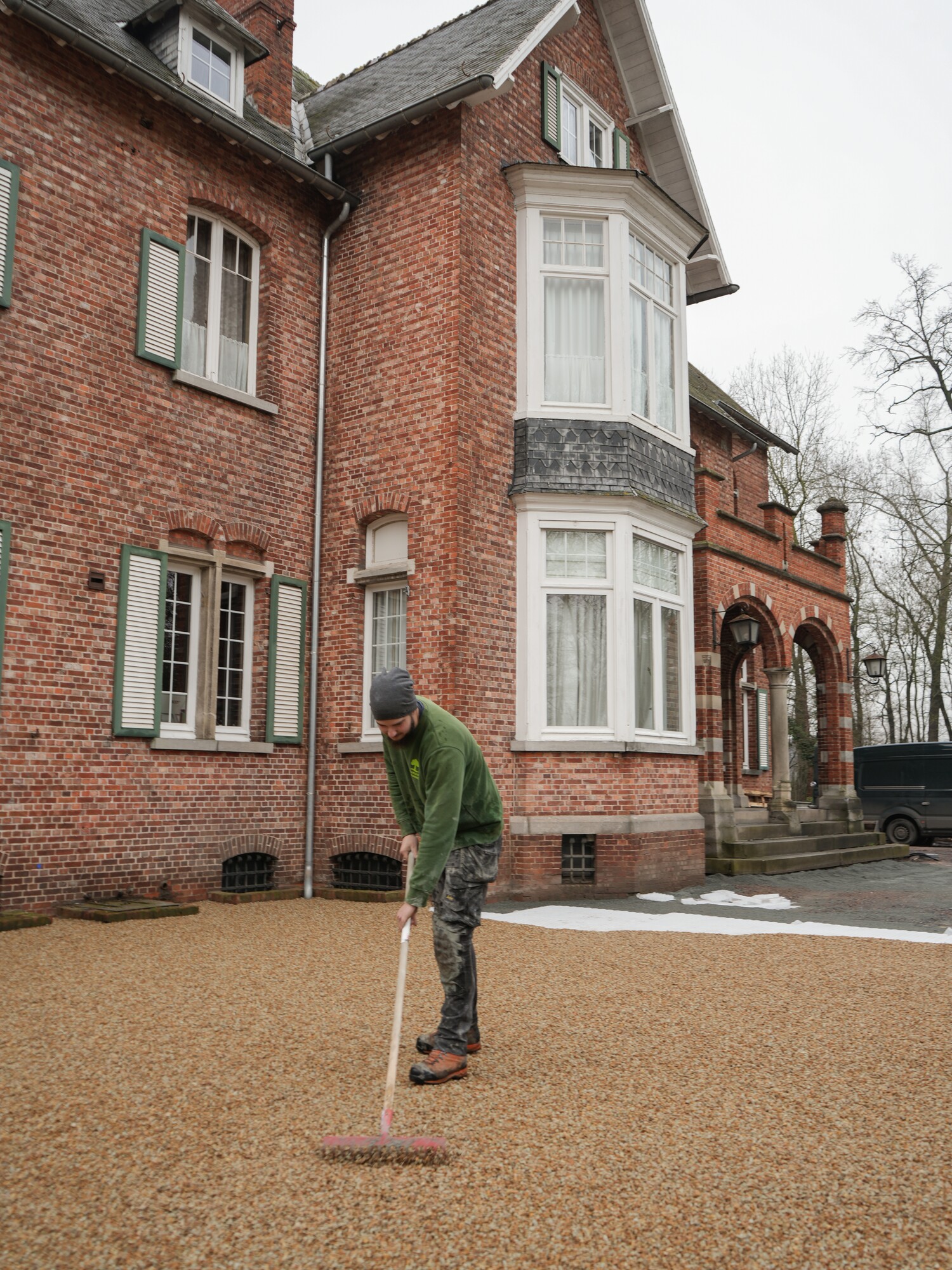 Man standing in a gravel driveway beside a large red-brick house on a cloudy day