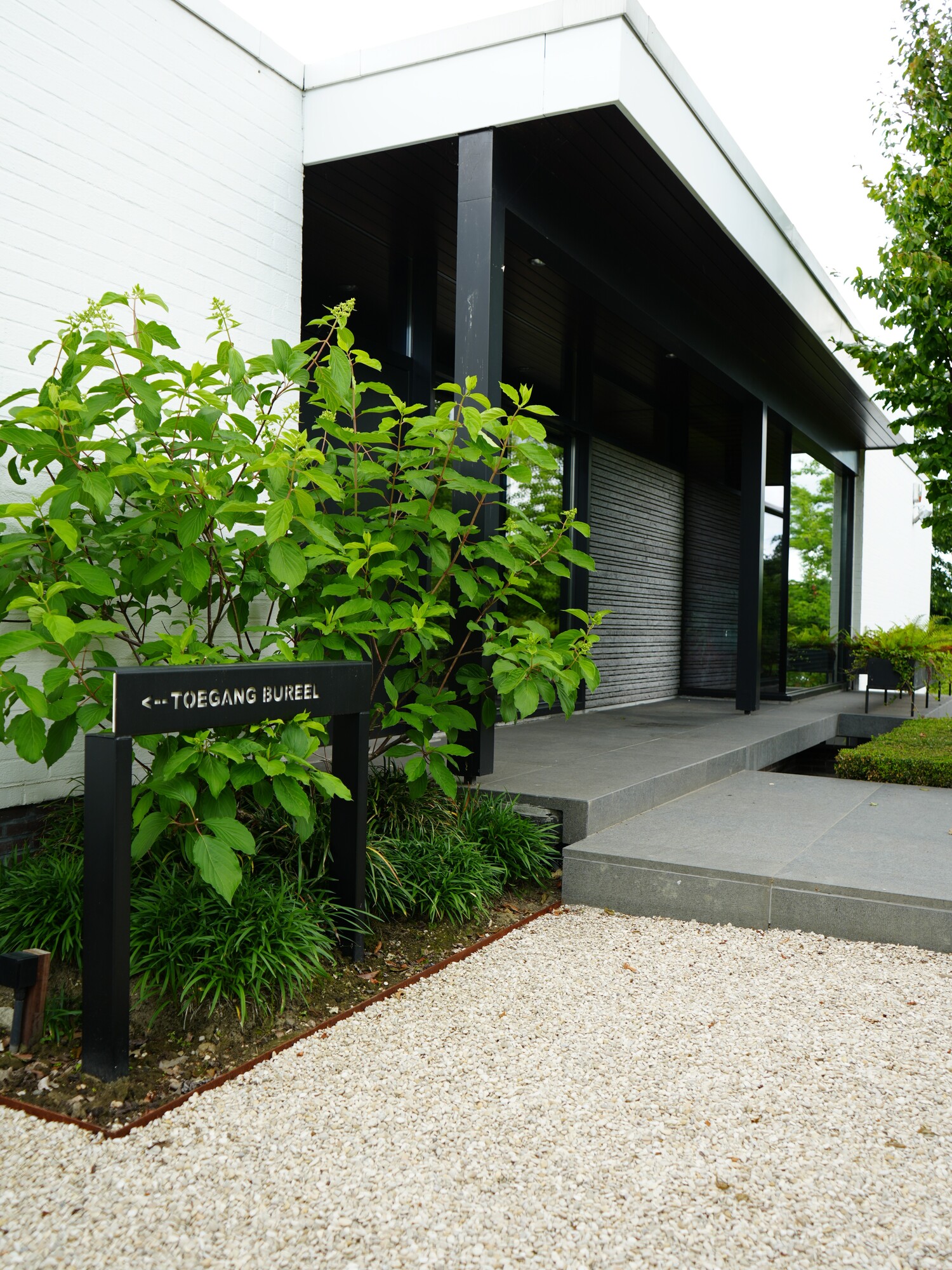 Entrée moderne d’un bâtiment en noir et blanc avec de la verdure et une allée en gravier