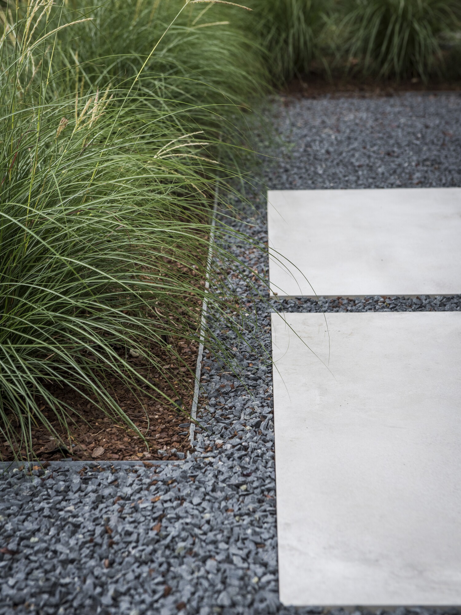 Betonnen stapstenen op een grindpad naast hoog groen gras