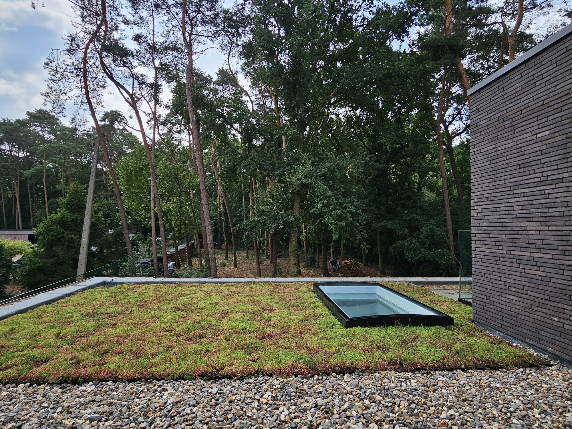 Gravel terrace next to a brick wall, overlooking a lawn with a flat roof skylight and trees.