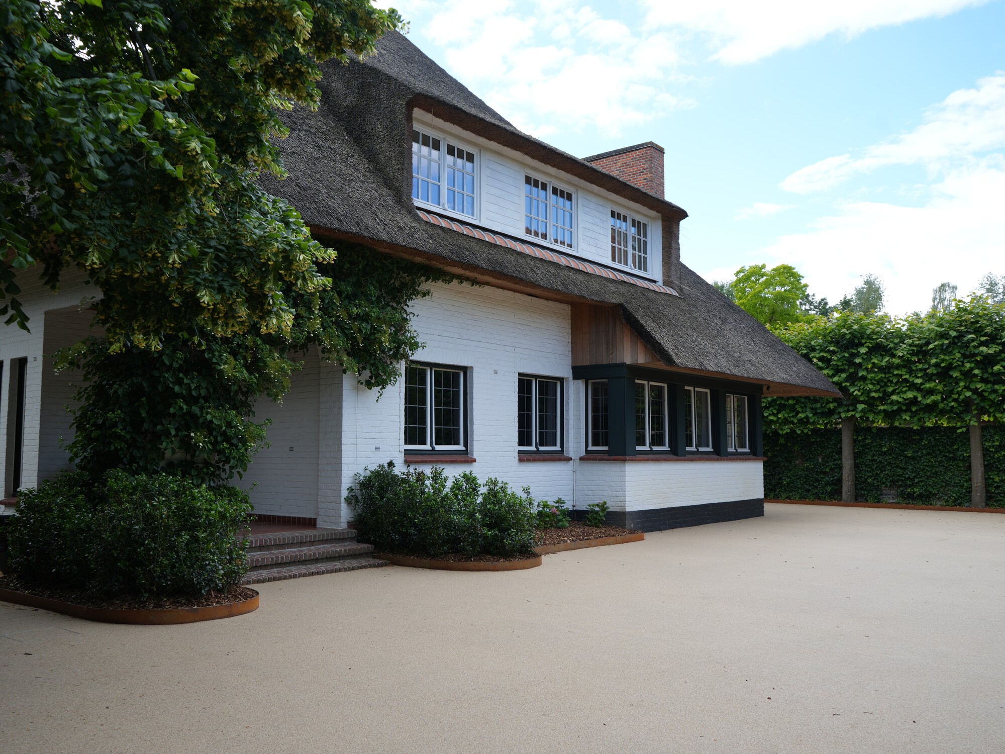 Two-story white house with dark roof, wide driveway, and hedges under blue sky
