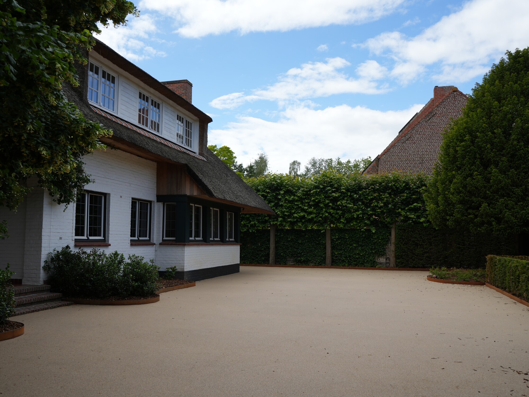 Wide paved courtyard beside a house, bordered by tall hedges under a blue sky