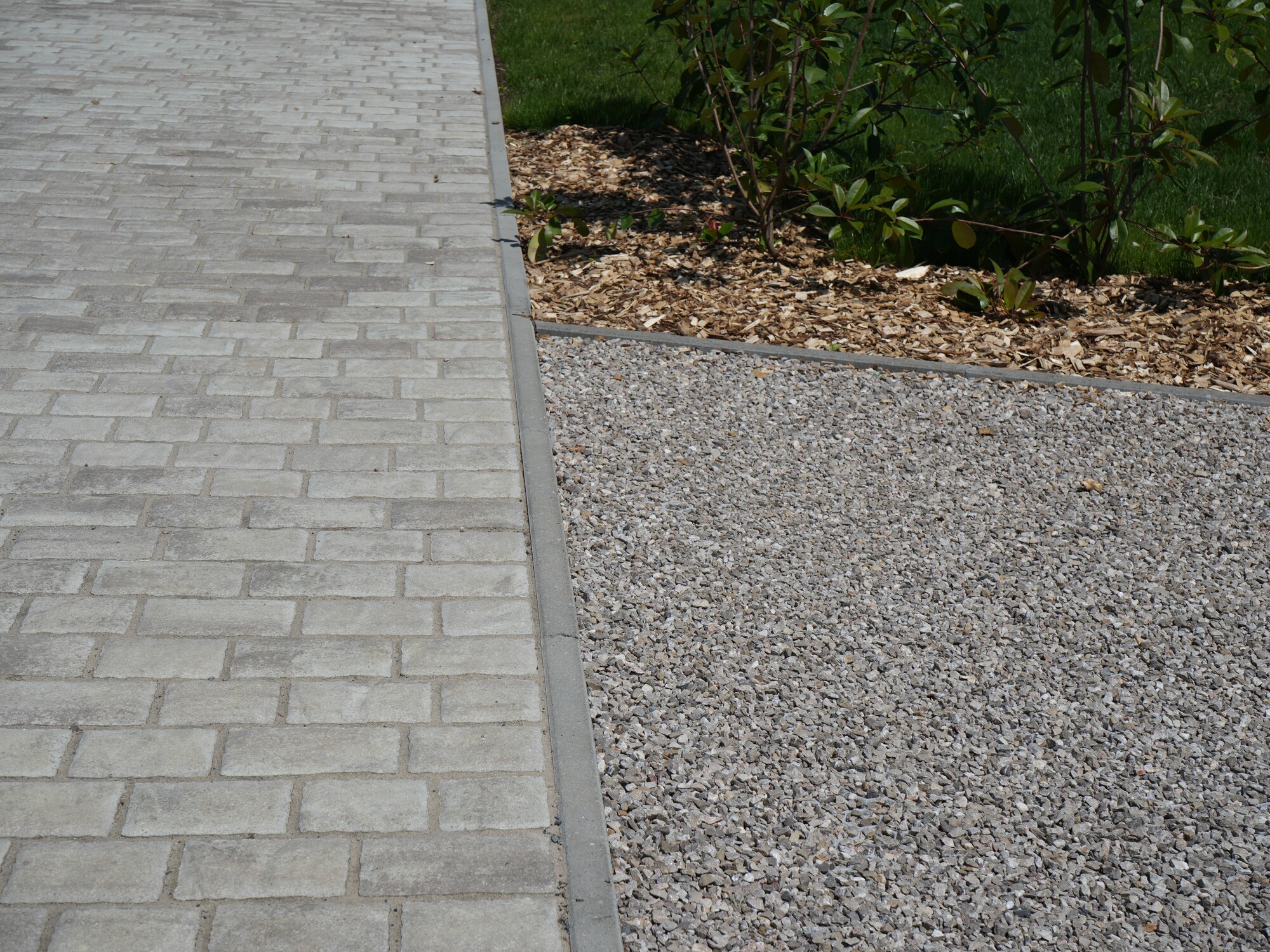 Paved walkway beside a gravel area and shrubs in sunlight