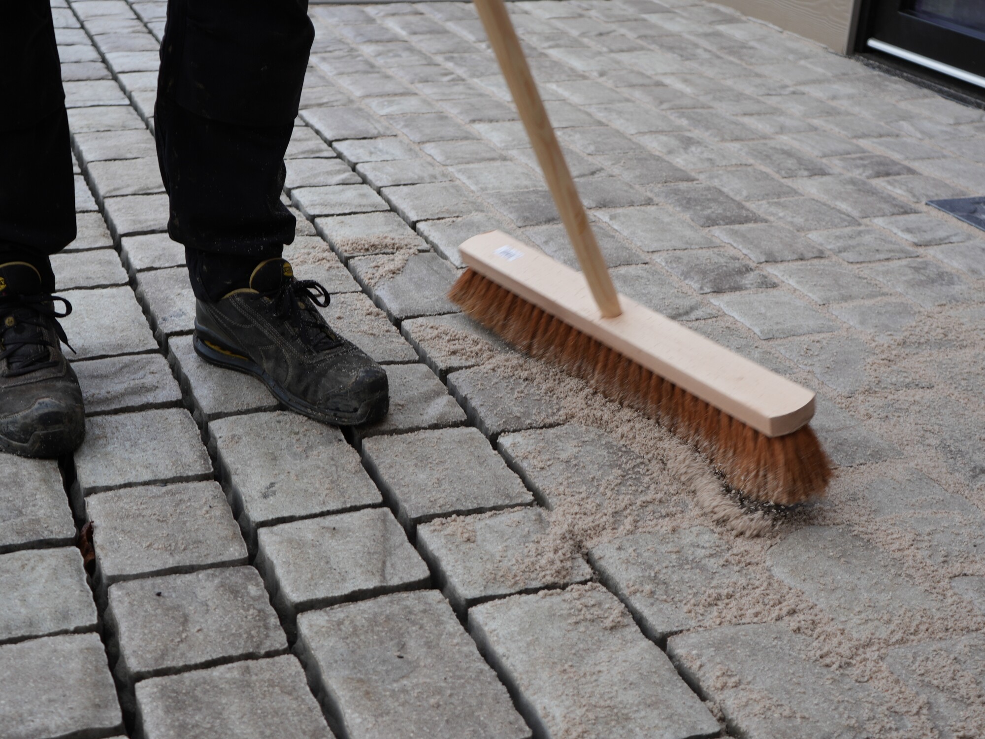 Person sweeping a cobblestone sidewalk with a push broom