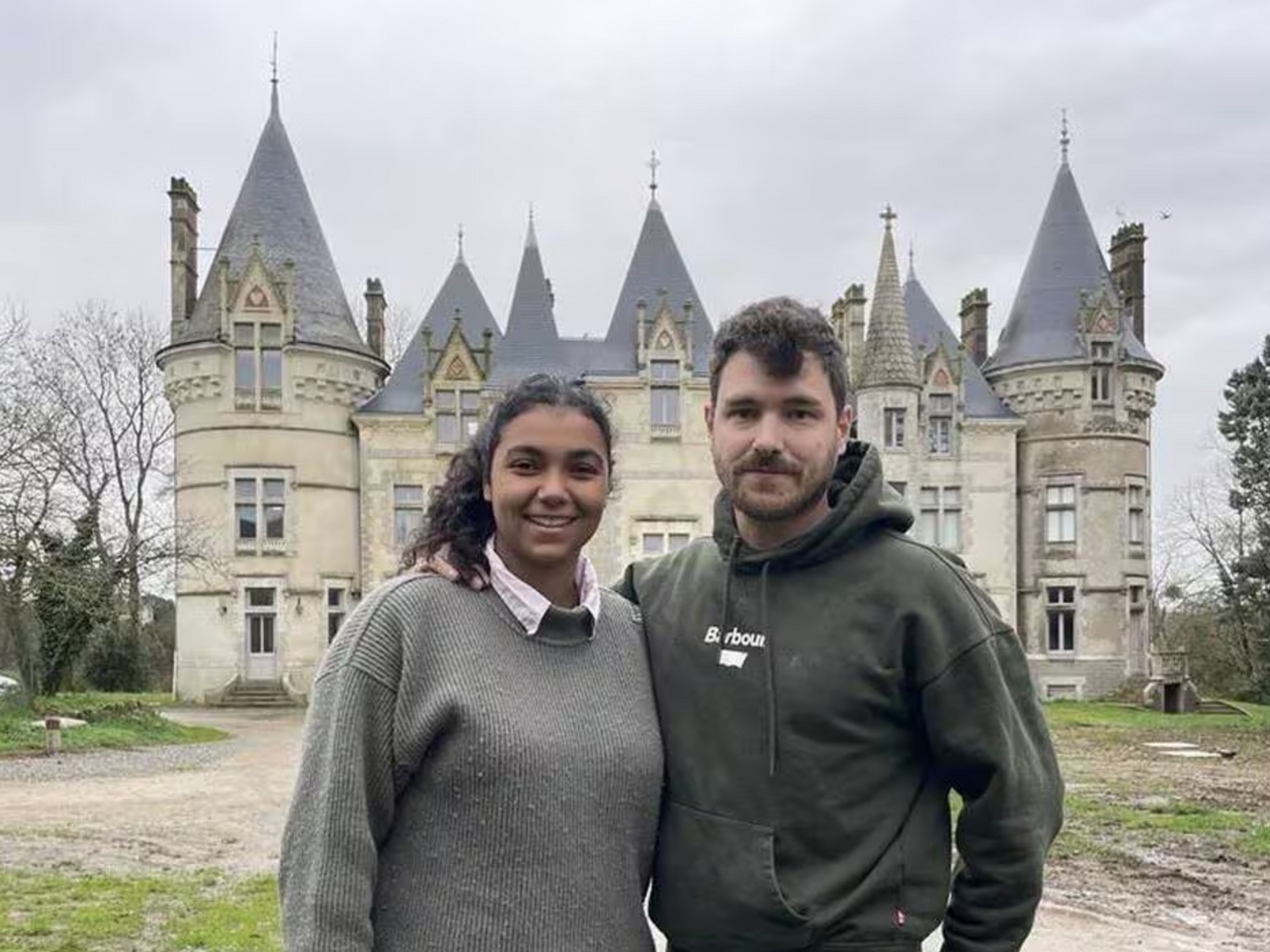 Deux personnes souriant devant un grand château en pierre par temps couvert.