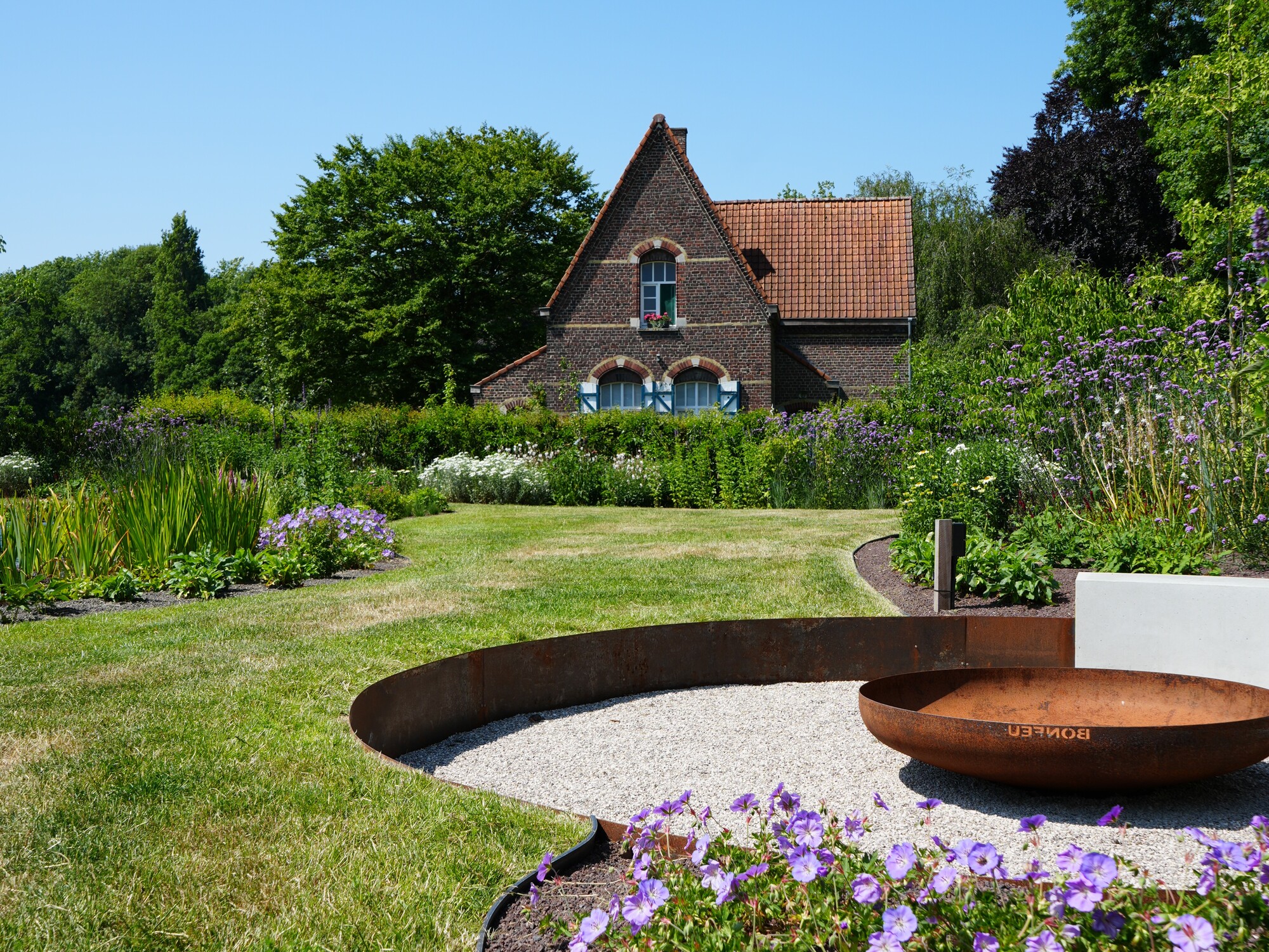 Curved garden beds with purple flowers in grassy yard, house and trees under blue sky