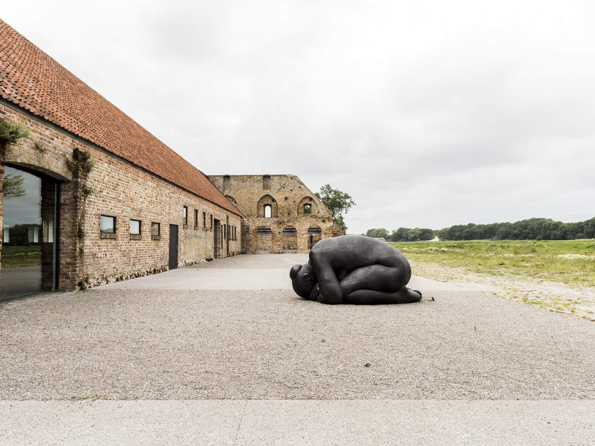 Large black sculpture on gravel beside a long brick building under cloudy sky