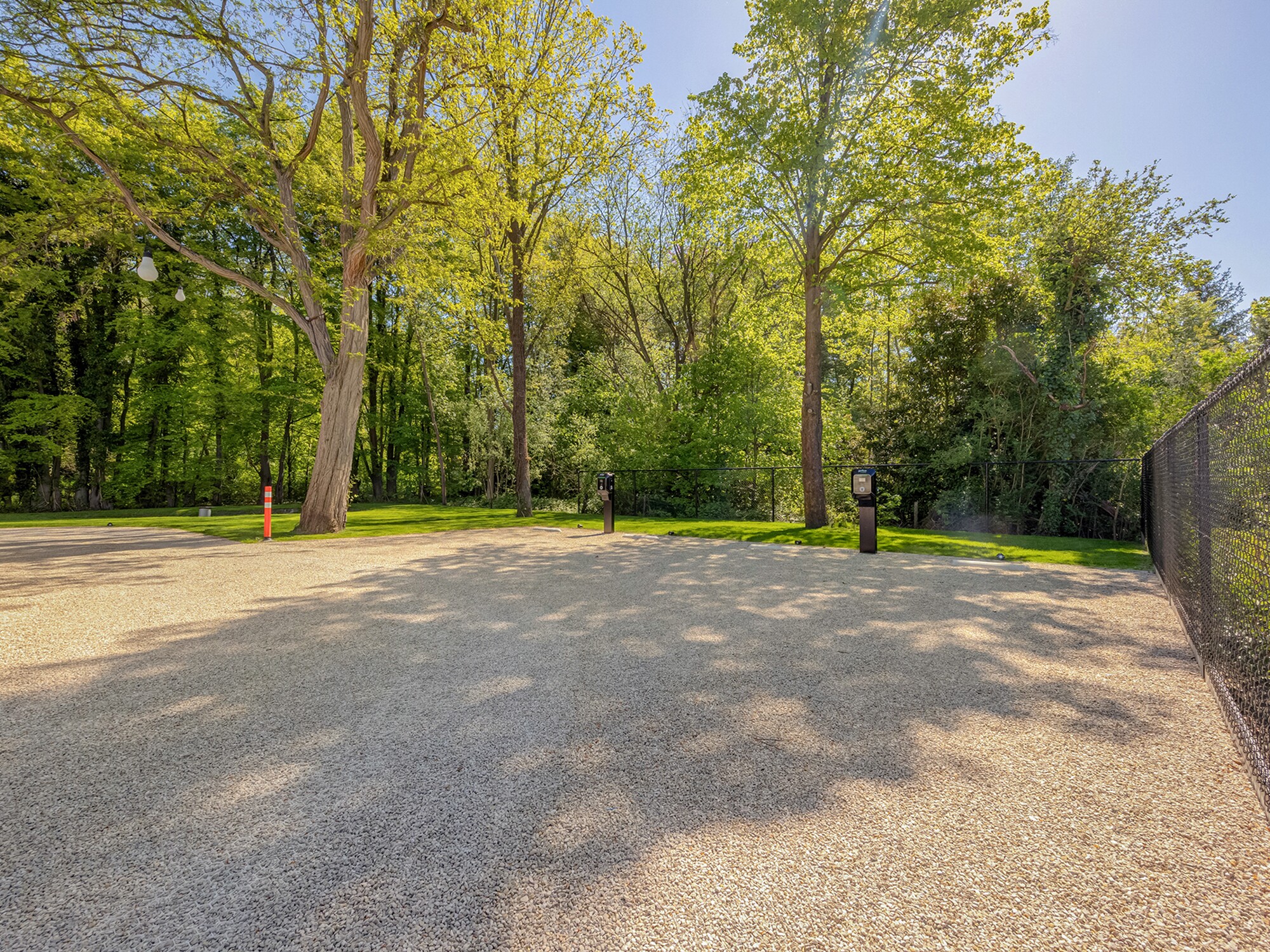 Sunlit paved courtyard beside a chain-link fence, surrounded by green trees