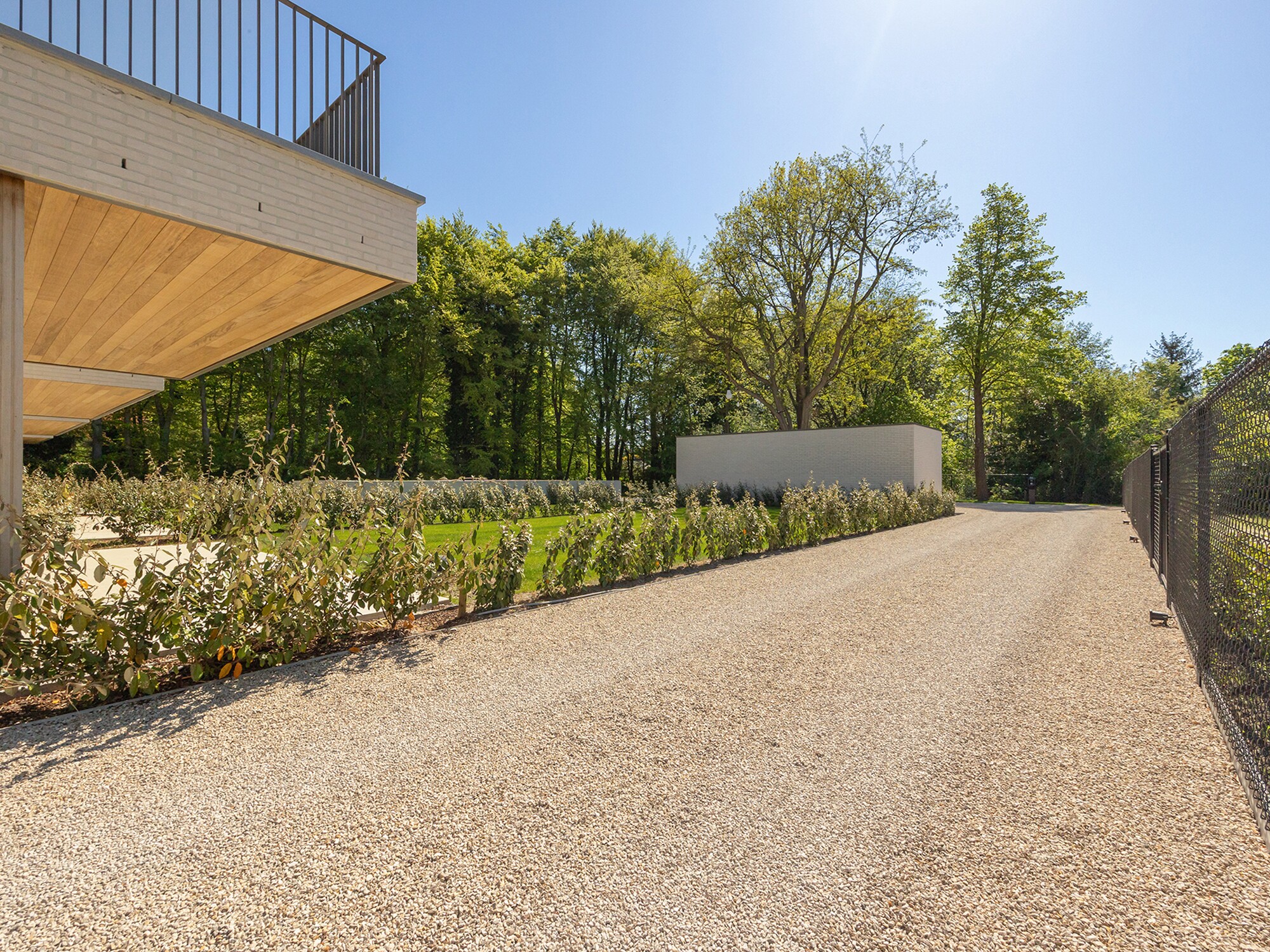 Sunlit gravel path beside modern building and fence, lined with shrubs and trees