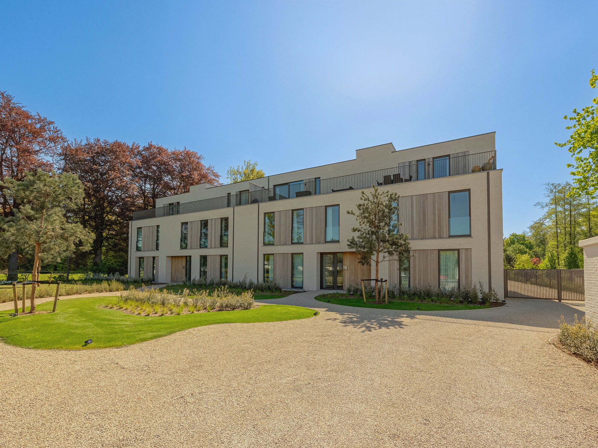 Modern white building with large windows, trees, and a gravel courtyard under blue sky