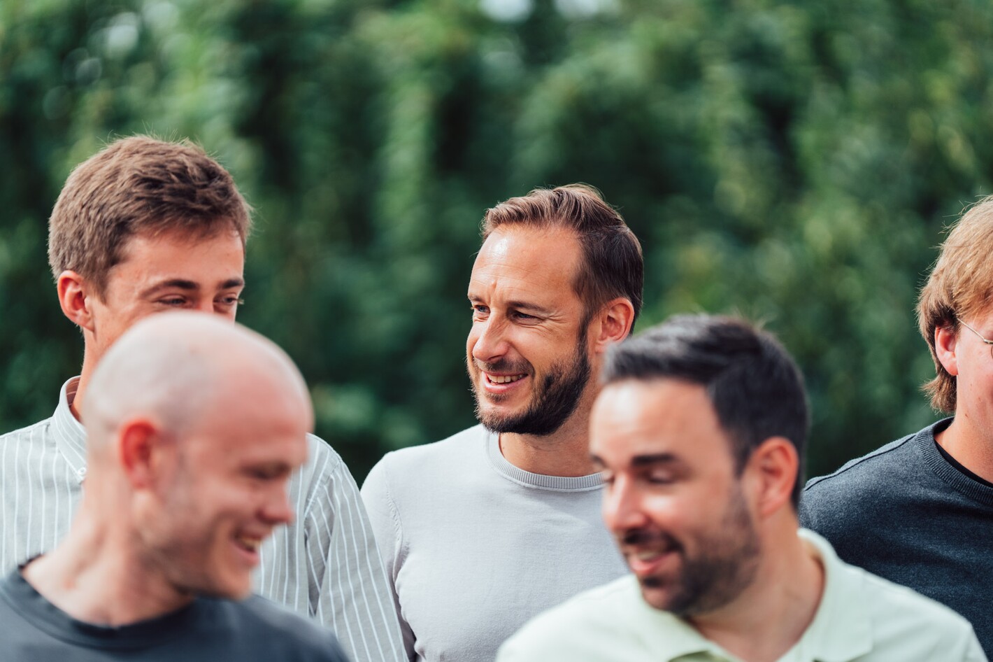 Groep mannen lacht samen buiten, met groen op de achtergrond.
